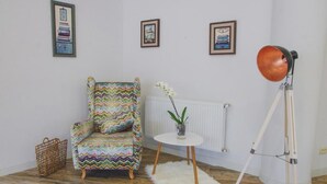 Ein Zimmer mit einem armchair im Zickzack-Muster, einem kleinen weißen Tisch mit einer Pflanze, einer Stehlampe und einem Heizkörper.