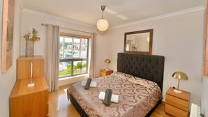 Un dormitorio con una cama grande, una mesita de noche, una lámpara, un espejo, una ventana con cortinas y vistas a la ciudad.