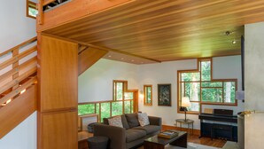 A spacious living area with a staircase, wooden flooring, a piano, and large windows offering a view of greenery. - Orcas Island, Cedar Sunrise #222 (Orcas Island)