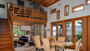 A spacious living area with a wooden staircase, a dining table with chairs, and a view of a lake through large glass doors. - Orcas Island, Cedar Sunrise #222 (Orcas Island)