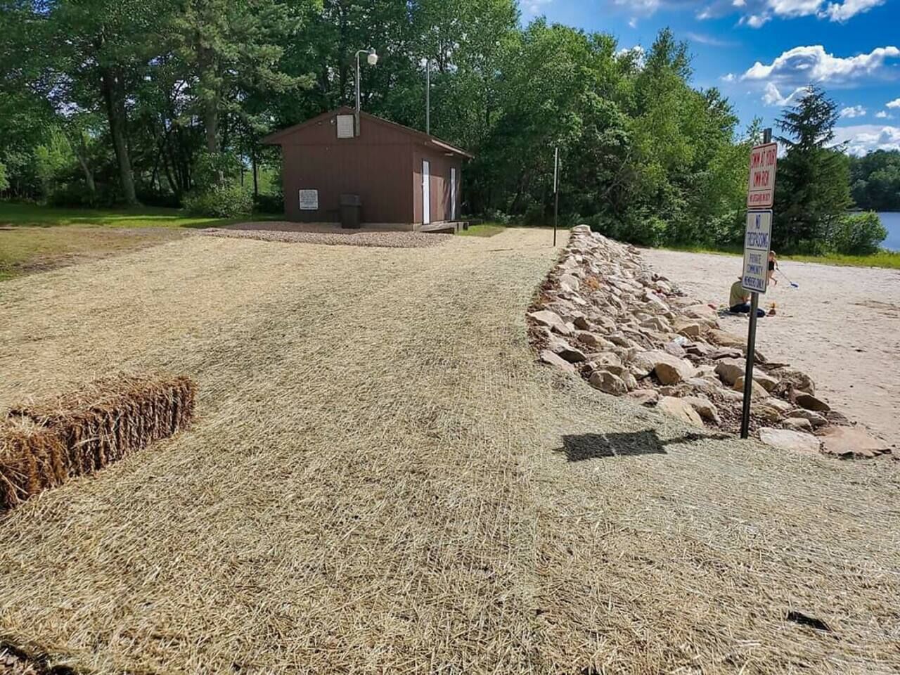 Un’area sterrata con un piccolo edificio in legno, un segnale stradale e un ambiente naturale con alberi e una distesa d’acqua sullo sfondo.