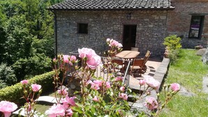 Un bâtiment en pierre avec un toit en tuiles, une terrasse avec une table et des chaises, et des fleurs roses au premier plan.