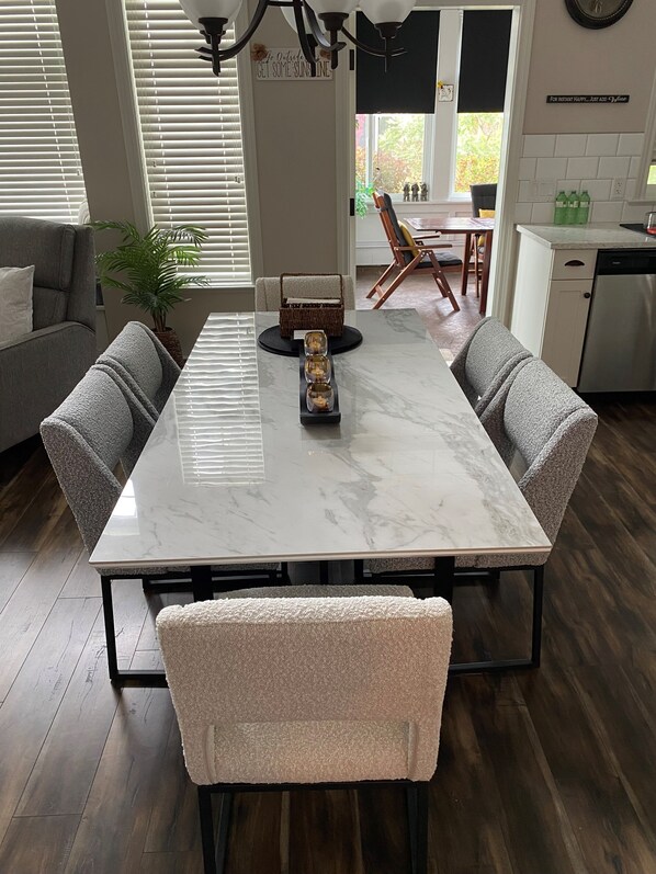 A modern dining area with a marble table, cushioned chairs, a potted plant, and a kitchen in the background. - Summer Short & Winter Long term rental at Canada's Warmest Lake Osoyoos (Oliver)
