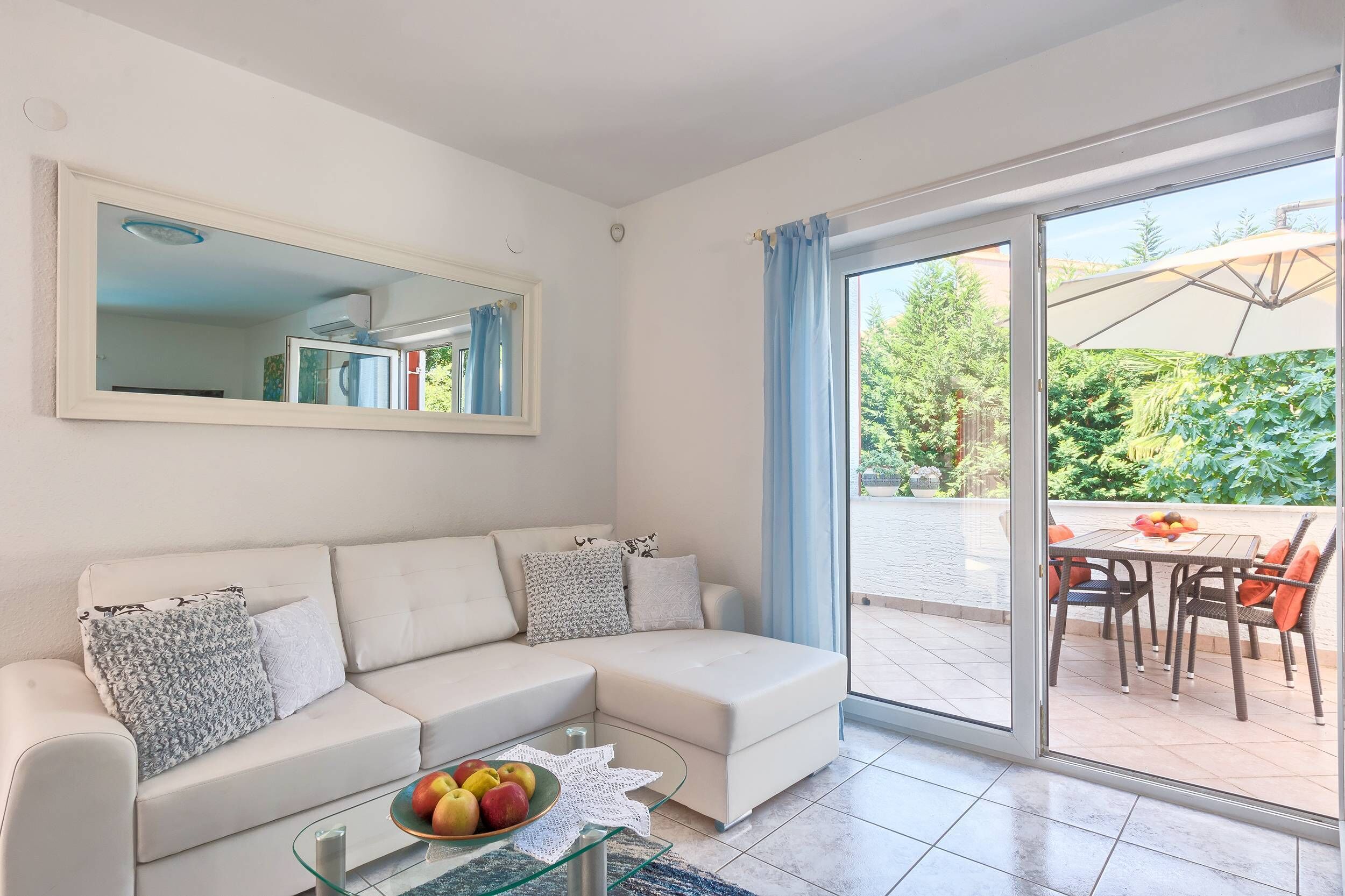 Un salon moderne avec un canapé blanc, une table basse en verre et une grande porte-fenêtre coulissante donnant sur une terrasse extérieure équipée d’une table et de chaises.