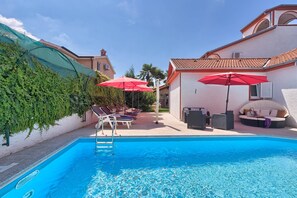 A swimming pool with lounge chairs and umbrellas, surrounded by a white building with a tiled roof and a covered area with greenery. - Villa Little Castle with private pool (Kukci)