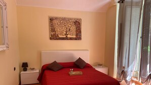 A bedroom with a red bedspread, a white headboard, and a decorative wall piece. There are two bedside tables with lamps and a small box labeled "Home."
