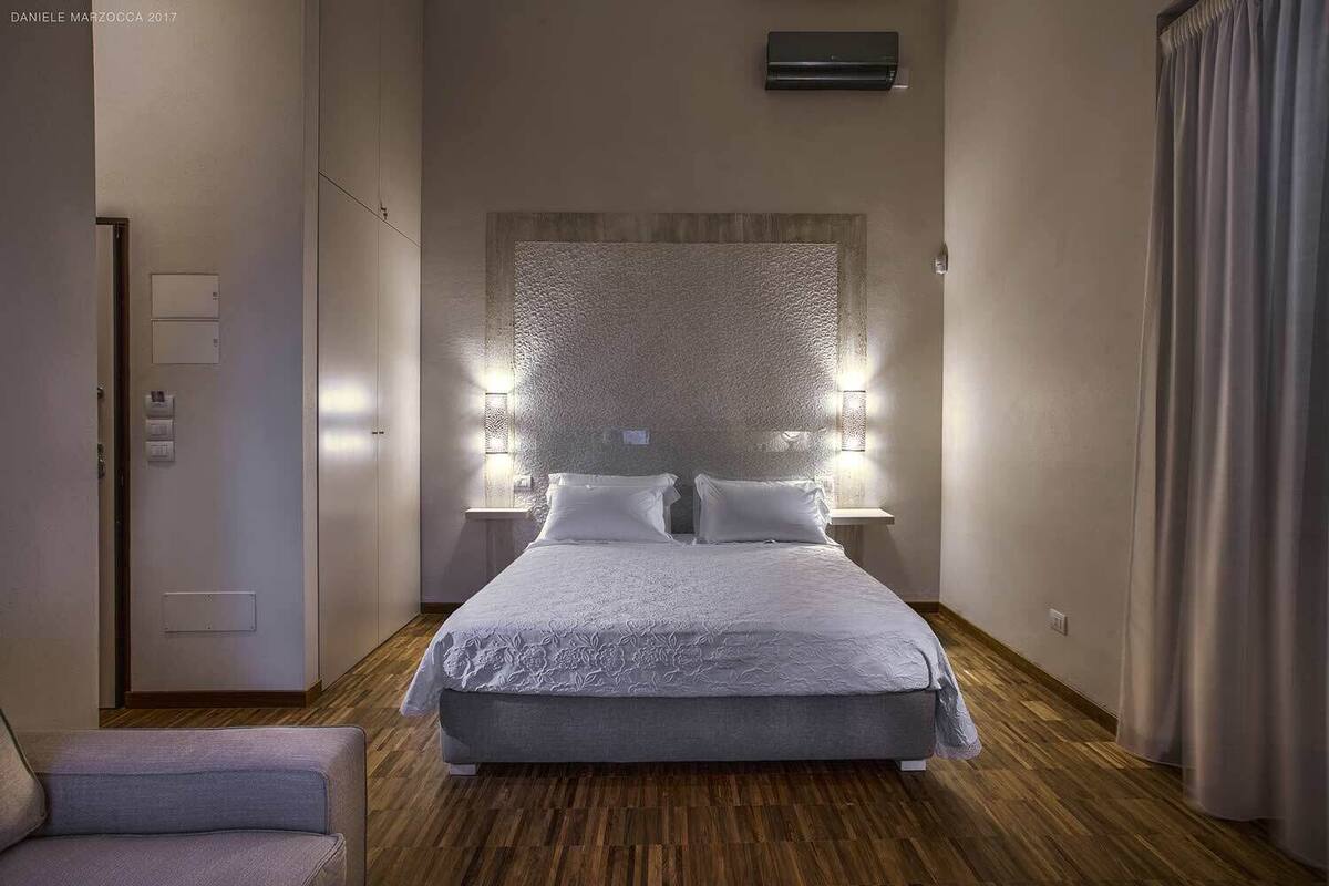 A modern hotel room with a large bed, a wooden floor, a wall-mounted air conditioner, and a small bedside table.
