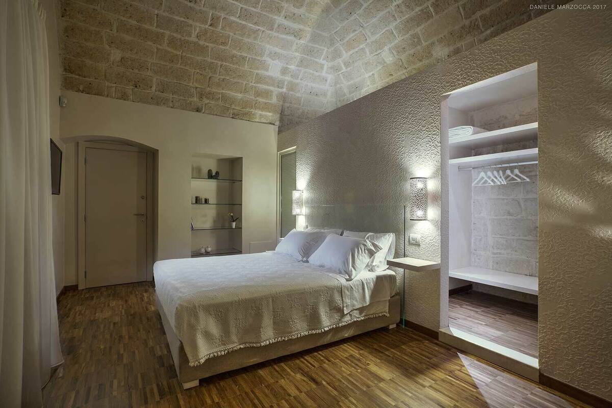 A bedroom with a wooden floor, a bed with white bedding, a built-in shelf unit, and a window with a view of a brick wall.