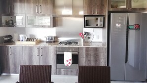 A modern kitchen with wooden cabinets, a stainless steel refrigerator, a microwave, and a gas stove. There are two wicker chairs and a toaster on the counter.