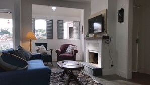 Ein Wohnzimmer mit blauem Sofa, einem grünen Sessel, einem gemusterten Teppich, einem Kamin und einem an der Wand befestigten Fernseher.