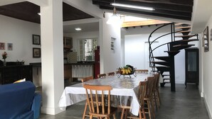 Ein Essbereich mit einem langen Tisch, Holzstühlen, einem blauen Sofa, einer Küche im Hintergrund und einer Wendeltreppe.