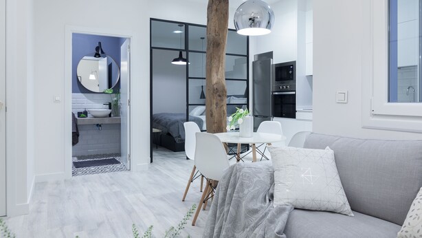 Un salon moderne avec un canapé gris, des chaises blanches, un tronc d’arbre en bois et une table à manger agrémentée d’un vase.