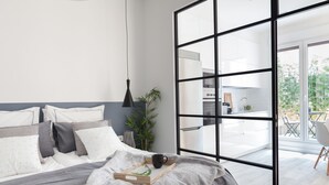 Une chambre moderne avec un grand lit, une table de chevet, une plante et une vue sur la cuisine et le balcon.