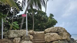 A private property entrance with a sign, a rope barrier, and a staircase leading up to a rocky area with palm trees in the background. - Beachfront-Chandler'sCoast-10mins to Holetown-2 BR apartment-private gated villa (Weston)
