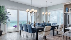 Un comedor con vista al océano, que incluye una mesa grande con sillas, iluminación colgante y una cocina moderna con refrigerador de acero inoxidable.