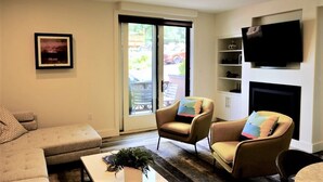 Ein Wohnzimmer mit einer Couch, zwei Sesseln, einem Couchtisch, einem Kamin, einem Fernseher und einer Glasschiebetür, die auf eine Terrasse führt.