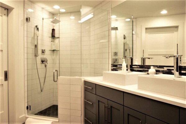 A modern bathroom with a glass-enclosed shower, white tiled walls, and a large mirror above the sink. - Olympic Terrace: Downtown Luxury & Convenience (Ketchum)