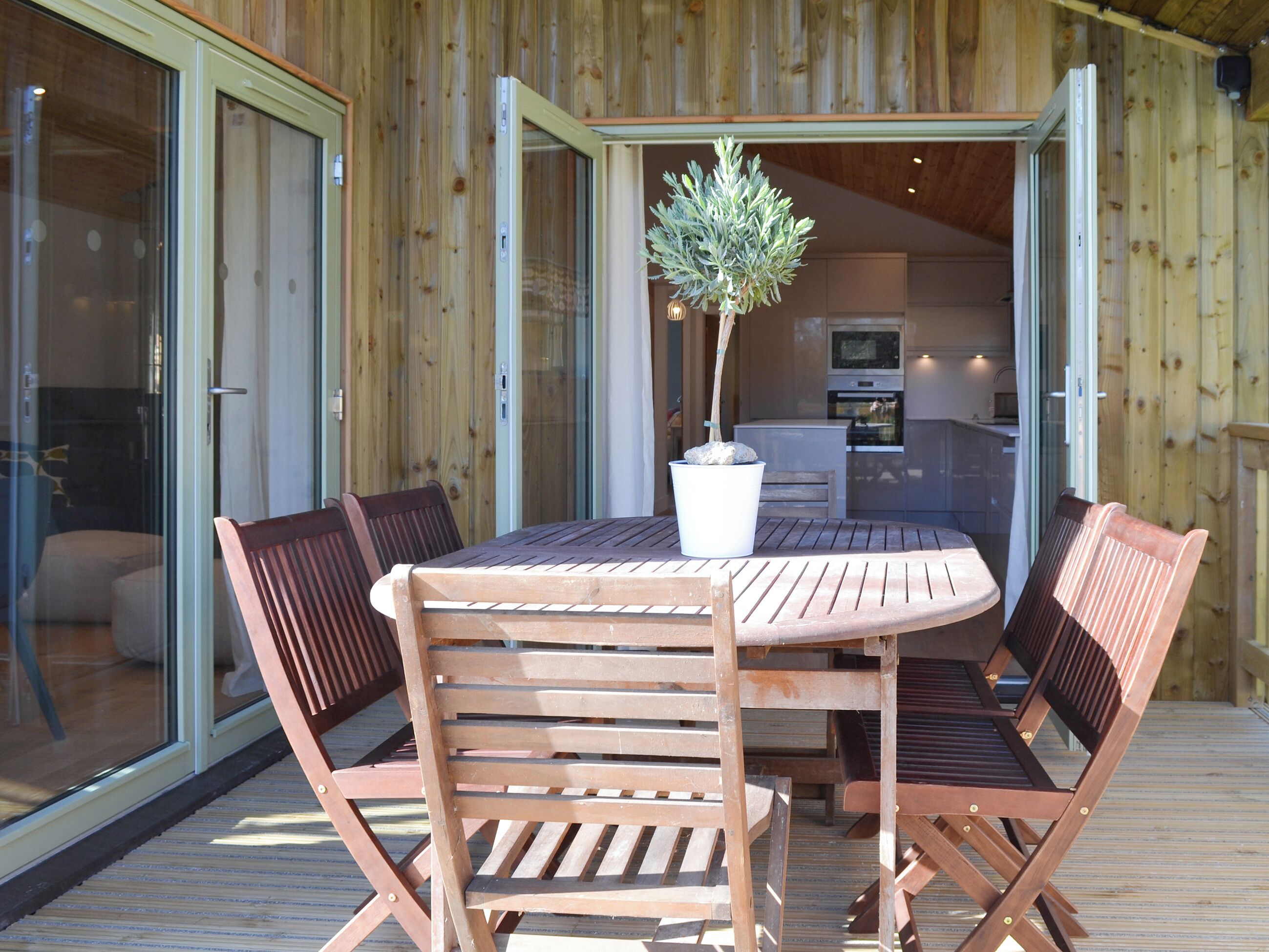 An open wooden deck with a round table and four chairs, a potted plant, and a view into a modern kitchen area.