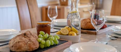 A breakfast setting with a loaf of bread, a bowl of grapes, lemons, a jar of jam, and a pitcher of water on a table.