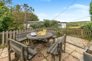 A wooden outdoor dining table with a tray of food and drinks, surrounded by a wooden fence and a garden. - 3 Bed in Lynton (oc-keepba) (Ilkerton)