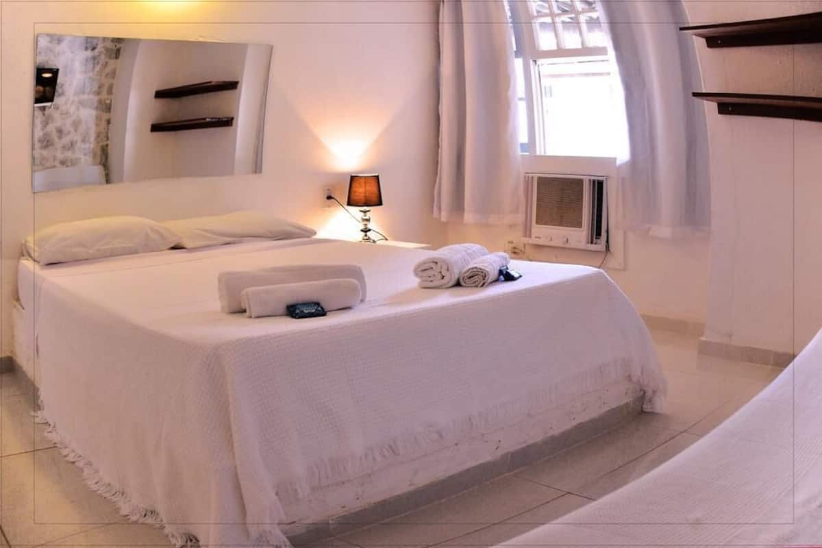 Una cama bien tendida con sábanas blancas, una lámpara de noche y un equipo de aire acondicionado instalado en la pared.