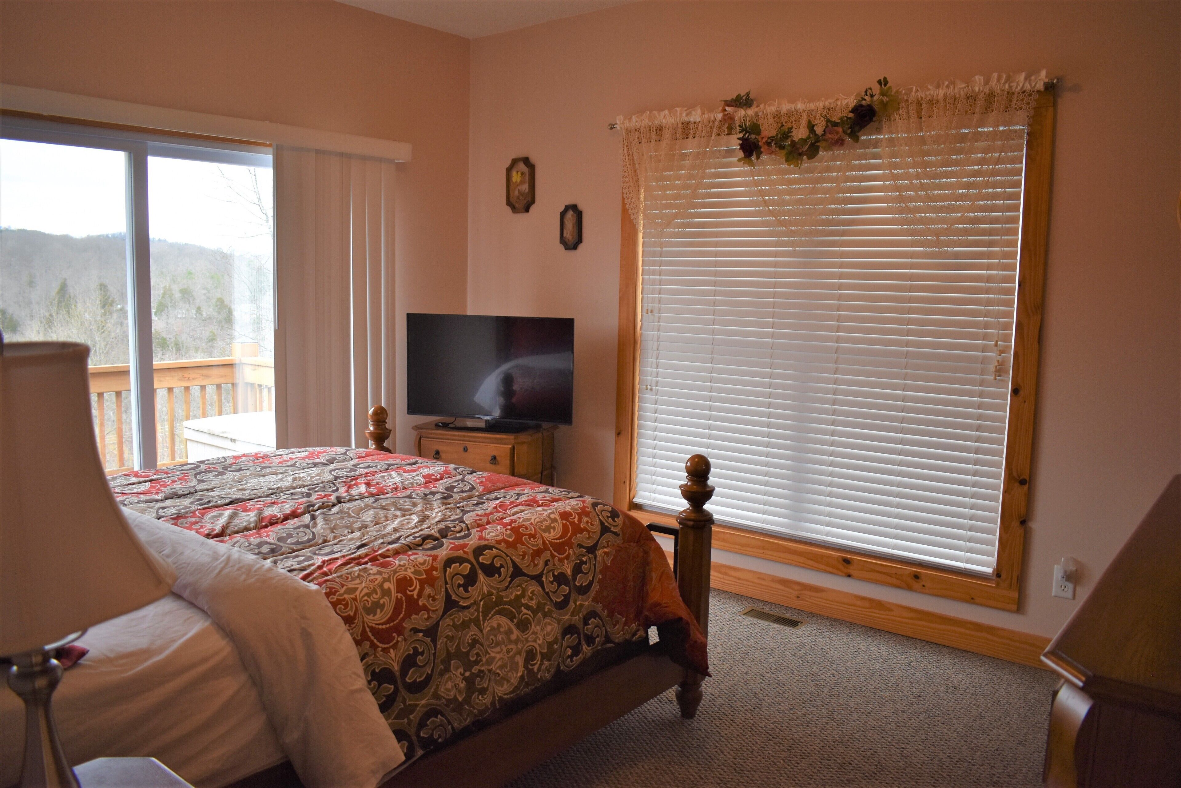 A bedroom with a bed, a TV, a window with blinds, and a view of the outdoors.
