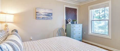 A bedroom with a bed, a dresser, a chair, a fan, a lamp, and a window with a view of trees.
