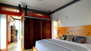 Un dormitorio con cabecera de madera, cama con colcha azul acolchada, suelo de madera, puerta de madera y una estantería con libros y una planta.