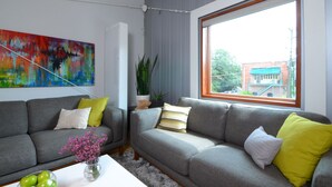 Una sala de estar moderna con dos sofás grises, cojines coloridos, un cuadro abstracto vibrante, un ventanal con vista a un edificio y una planta en maceta.