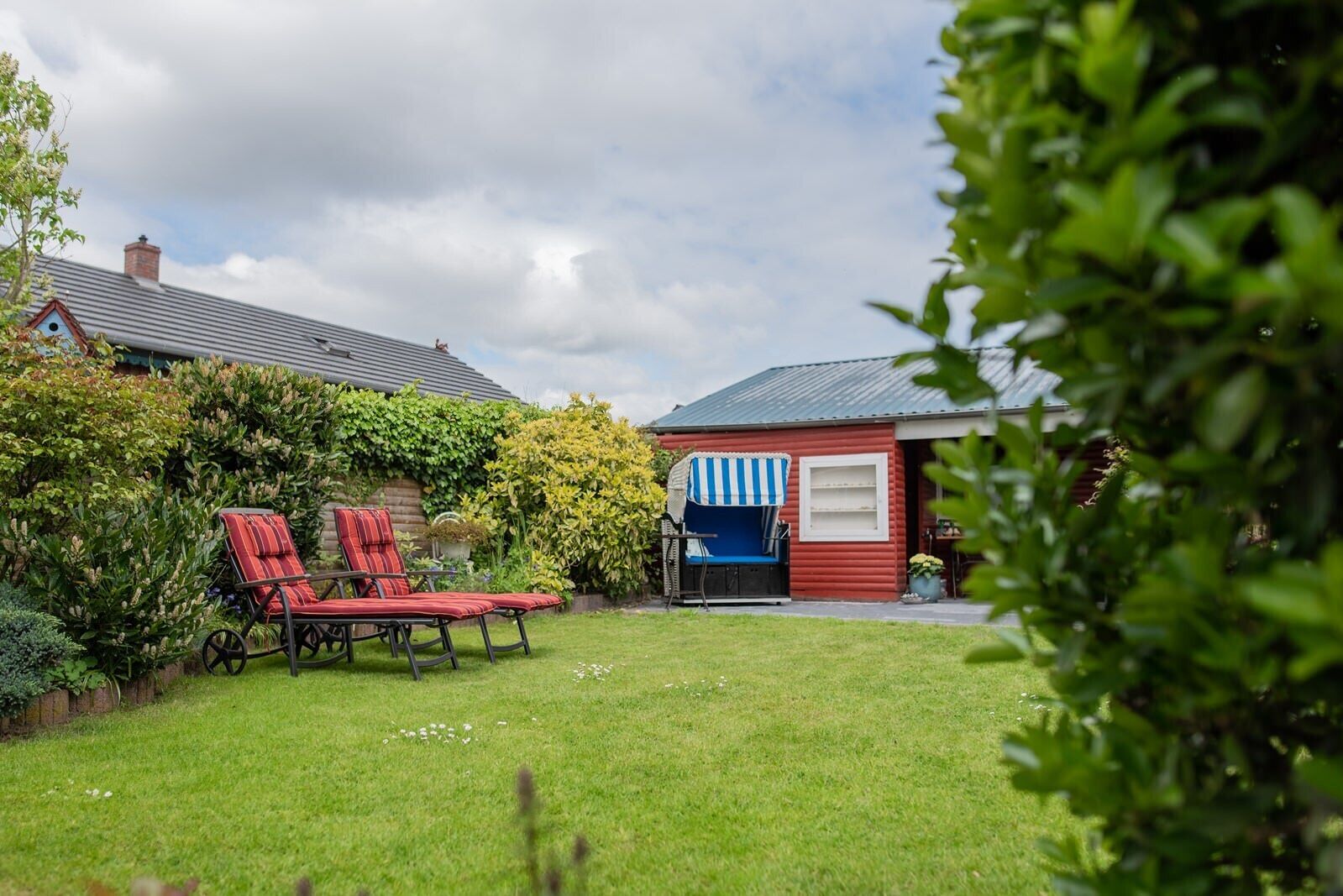 Ein Garten mit einem roten Hollschuppen, einer blau-weißen Markise und zwei roten Liegestühlen.