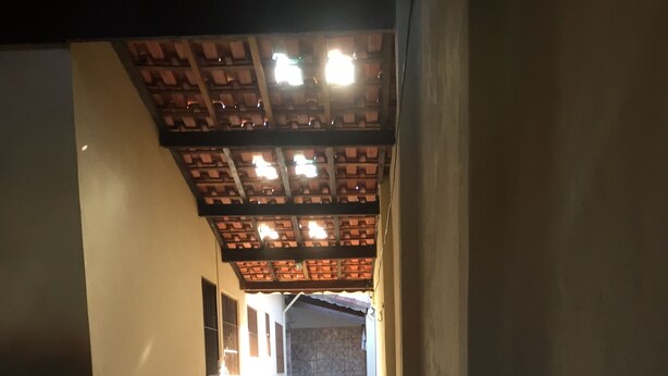 Un couloir au sol carrelé, un plafond avec des poutres apparentes et une vue sur une pièce dont le mur est en pierre et qui contient une table.