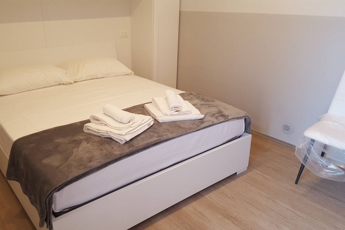 A neatly made bed with white linens and a gray blanket, a small bedside table with a white towel, and a wooden floor.