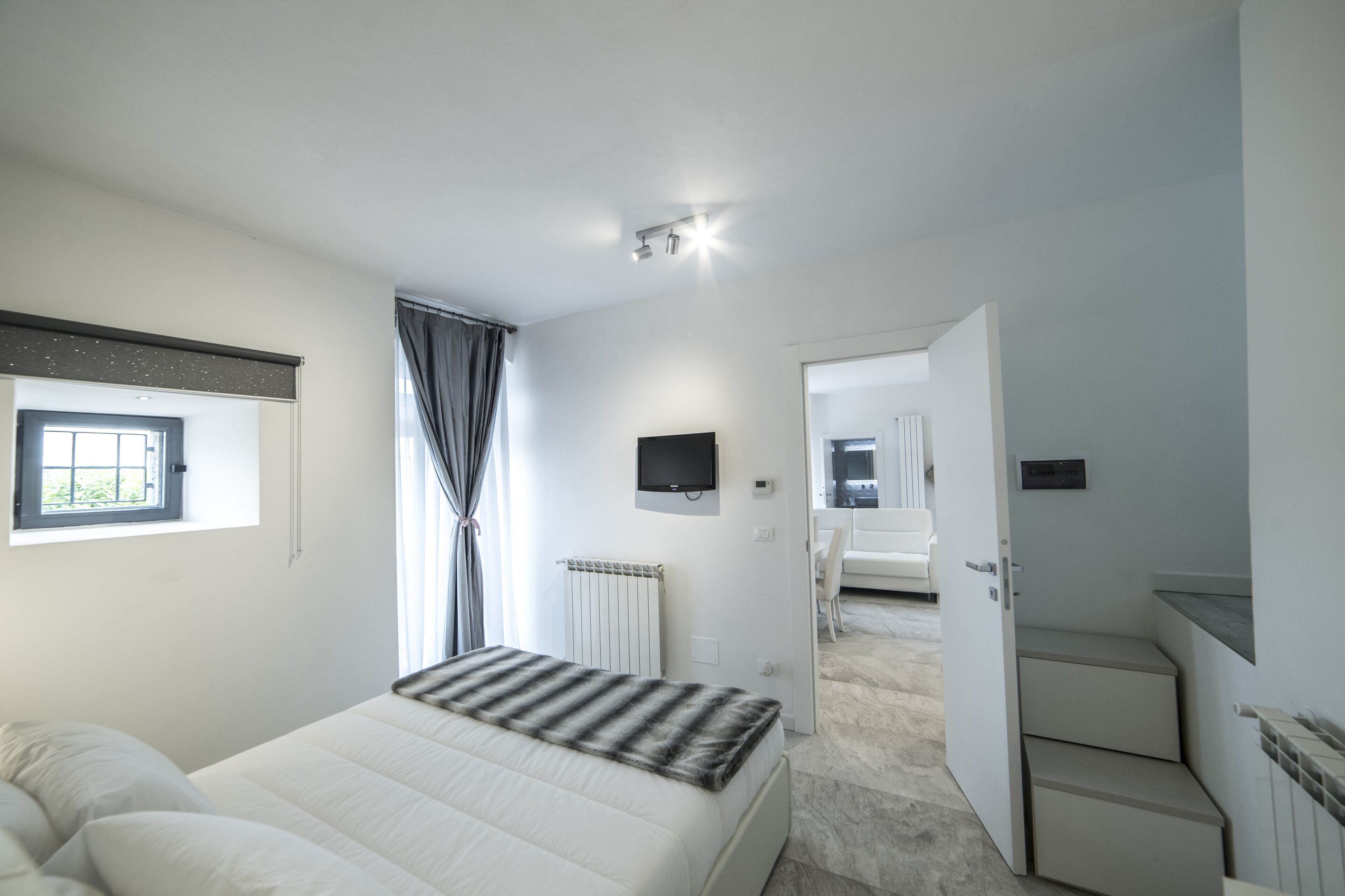 A modern bedroom with a bed, a TV, a window with curtains, a radiator, and a staircase leading to a bathroom.