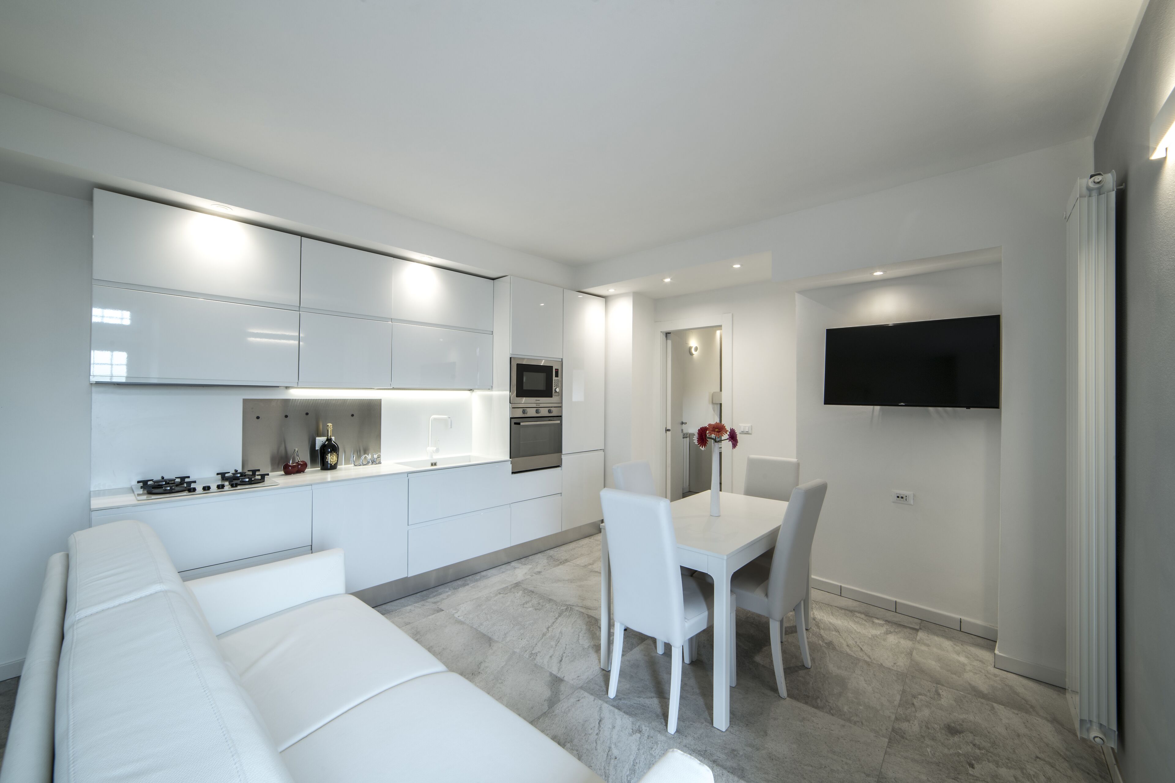 Una cocina moderna con armarios blancos, una zona de comedor con mesa y sillas blancas, y un televisor de pantalla plana montado en la pared.