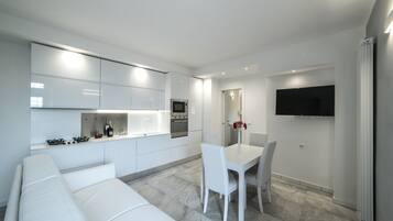 Una cocina moderna con armarios blancos, una zona de comedor con mesa y sillas blancas, y un televisor de pantalla plana montado en la pared.