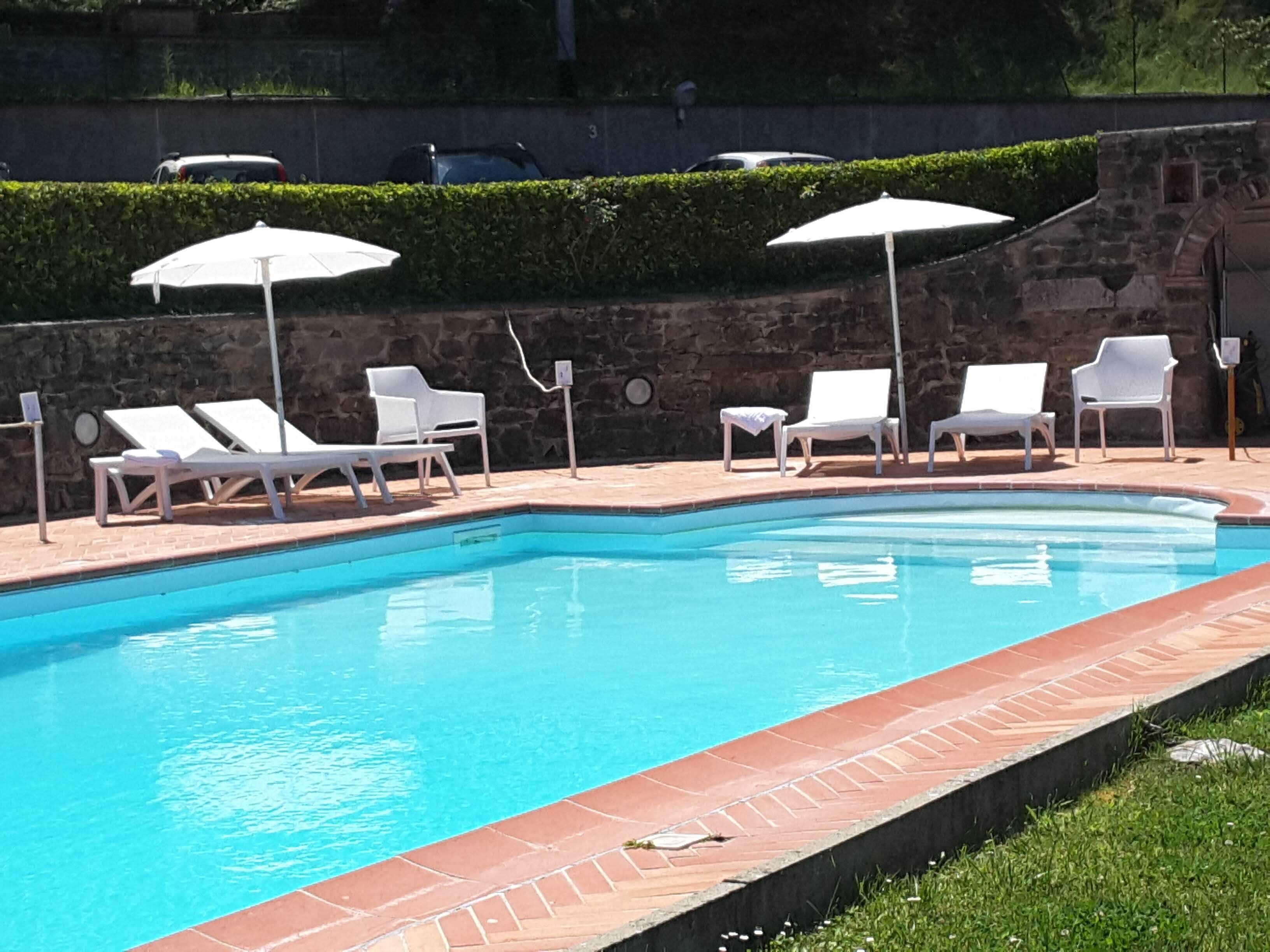 Piscina con sillas de descanso blancas y sombrillas, rodeada de muros de piedra y follaje.