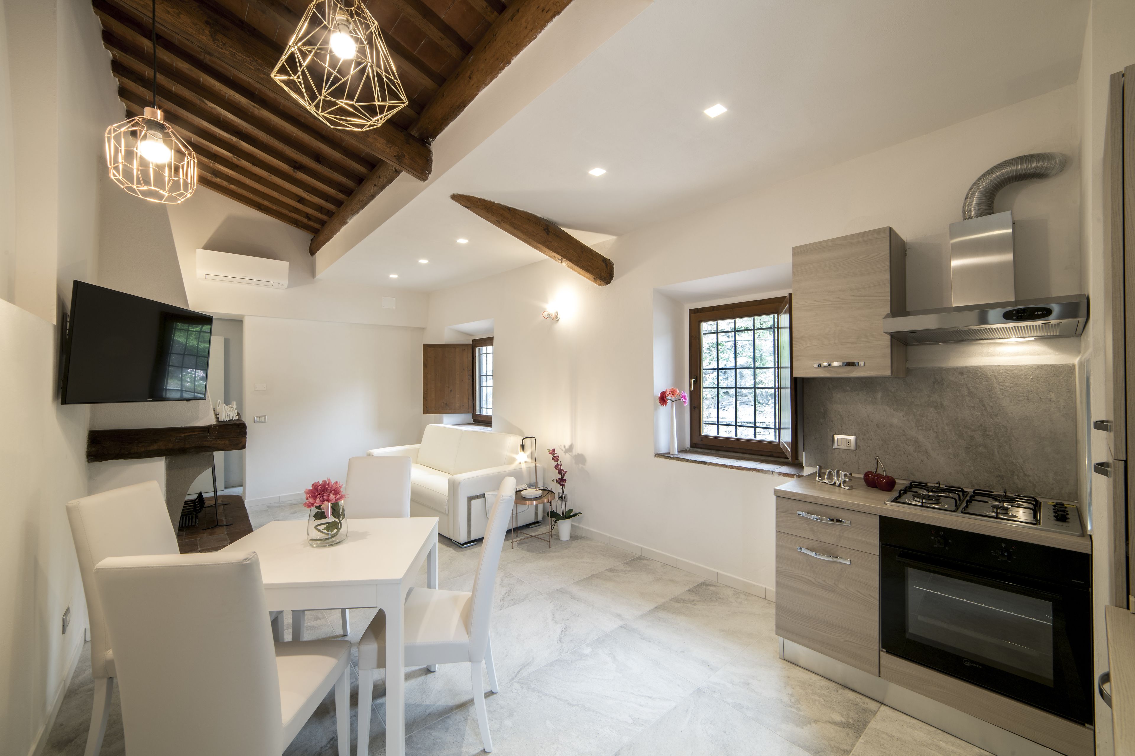 Una cucina moderna con forno integrato, piano cottura a gas e cappa aspirante in acciaio inossidabile. C'è una zona pranzo con tavolo e sedie bianche, e un televisore montato a parete.