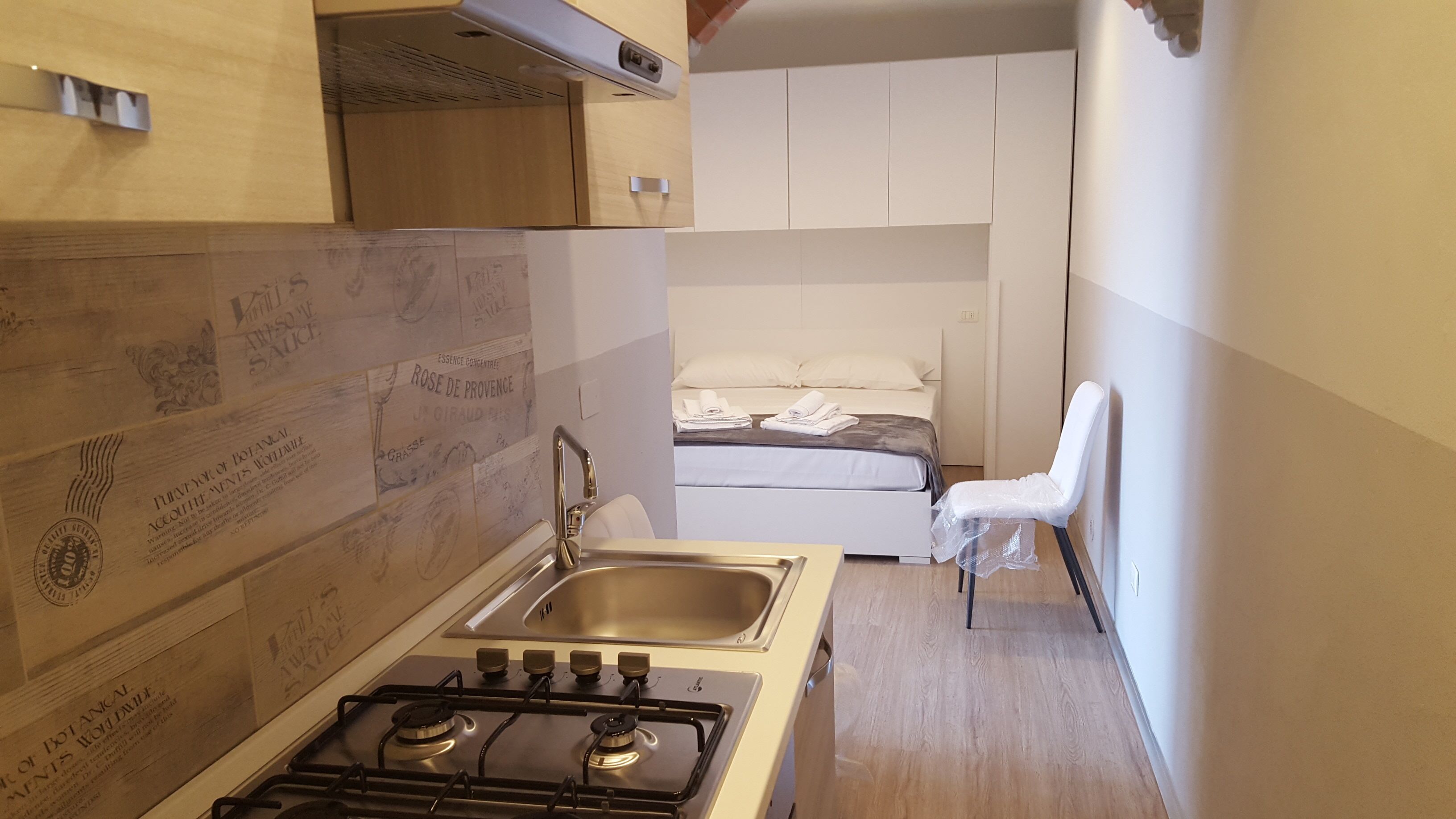 A compact kitchen with a gas stove, sink, and wooden backsplash. A small bed with a mattress and pillows is visible in the background.
