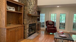 A living room with a stone fireplace, wooden shelving, a leather armchair, and large windows.