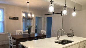 A modern kitchen with a white countertop, stainless steel sink, and a dishwasher. There is a dining table with chairs and a chandelier above.
