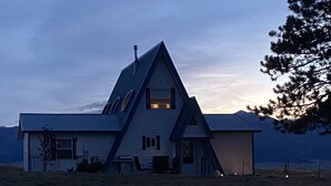 Une grande maison moderne, dotée d’un toit à forte pente et de nombreuses fenêtres, entourée d’un paysage verdoyant et avec une montagne en arrière-plan.