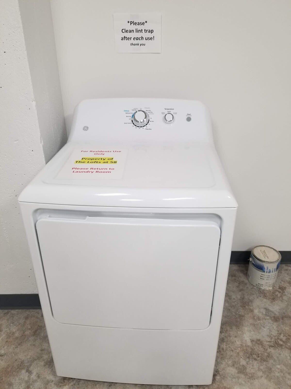 A front-loading washing machine with a control panel, a sign requesting to clean lint traps, and a note for residents to return to the laundry room.