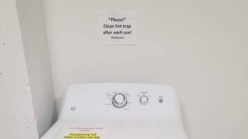 A front-loading washing machine with a control panel, a sign requesting to clean lint traps, and a note for residents to return to the laundry room.