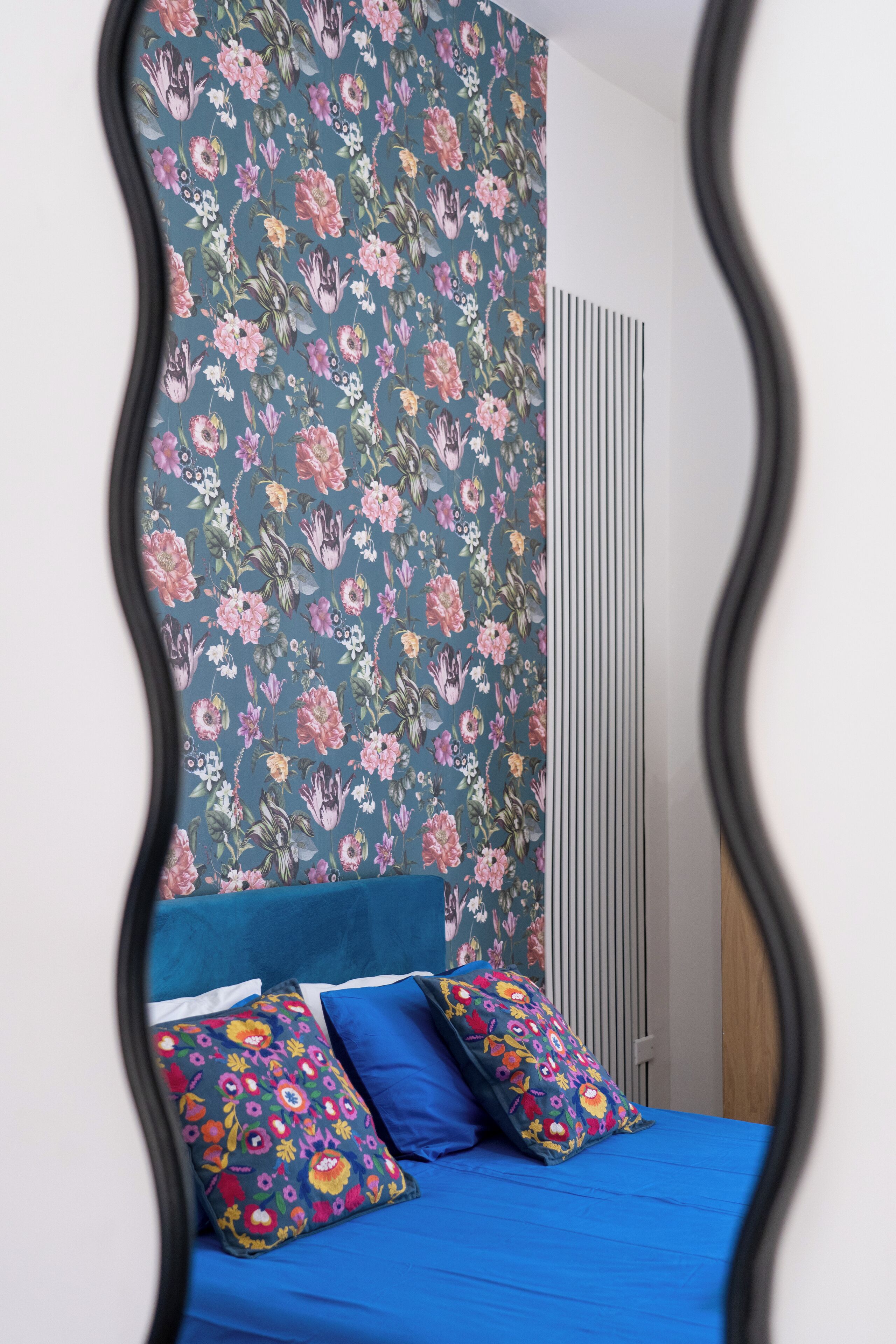 A bedroom with a floral wallpaper, a blue bedspread, and colorful pillows.