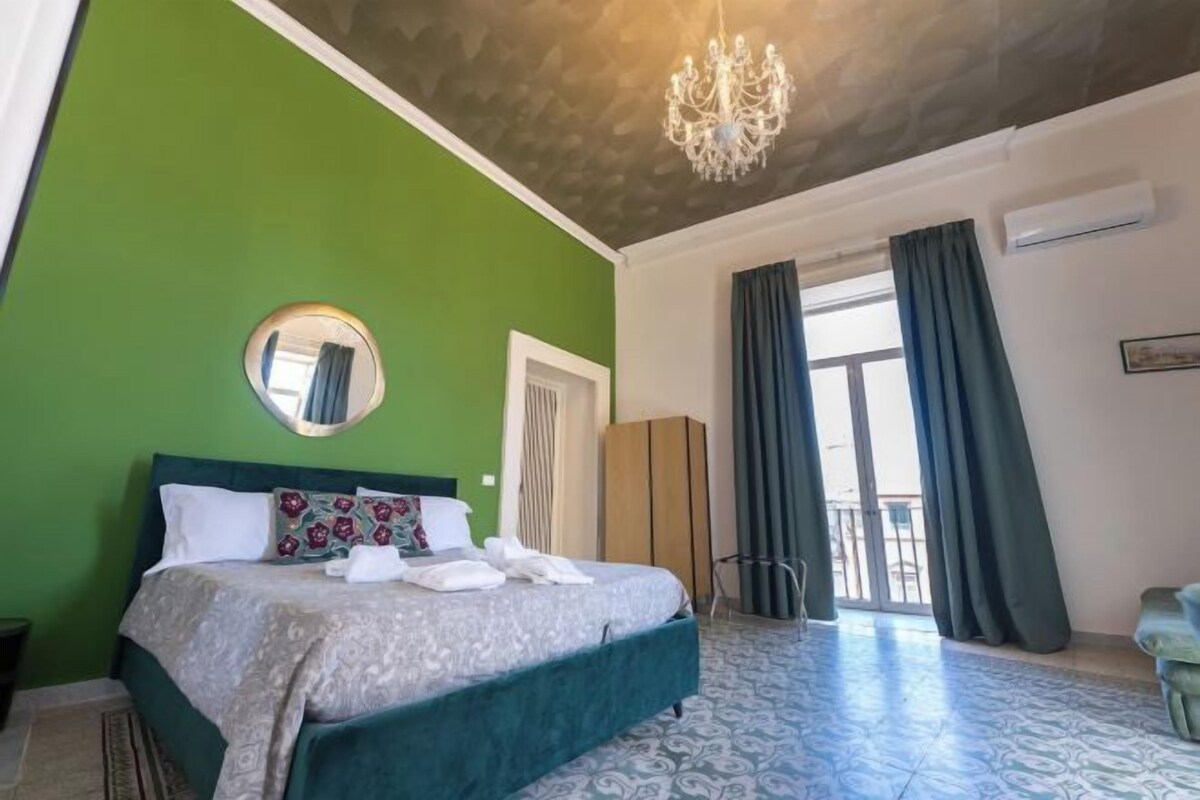 A hotel room with a green wall, a large bed with a patterned bedspread, a chandelier, a view of the outdoors through a sliding door, and a small seating area with a green sofa.