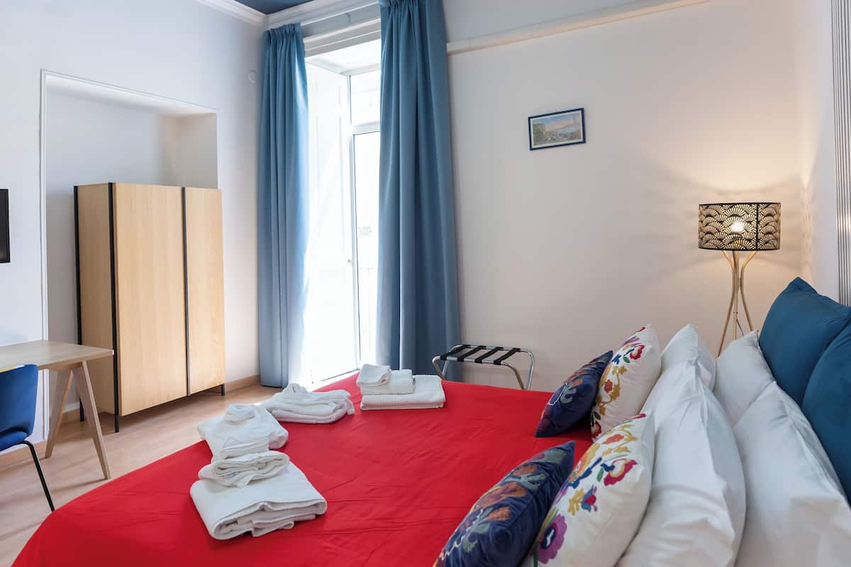 Een slaapkamer met een rode spreideken, blauwe gordijnen, een houten garderobe en een bureau.