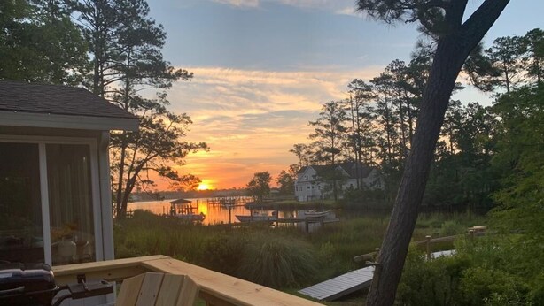 A wooden deck with a grill and a chair, overlooking a sunset over a lake with houses in the background.
