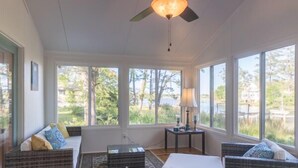 A sunlit room with wicker furniture, a ceiling fan, and large windows offering a view of greenery and a body of water.