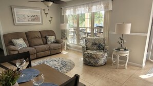 Una sala de estar con un sofá marrón, un sillón estampado, una mesa de comedor con individuales y un ventilador de techo.