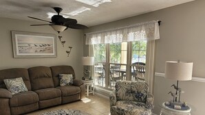 Un salón con sofá marrón, sillón estampado, alfombra redonda, ventilador de techo y vistas a un patio exterior con sillas y mesas.